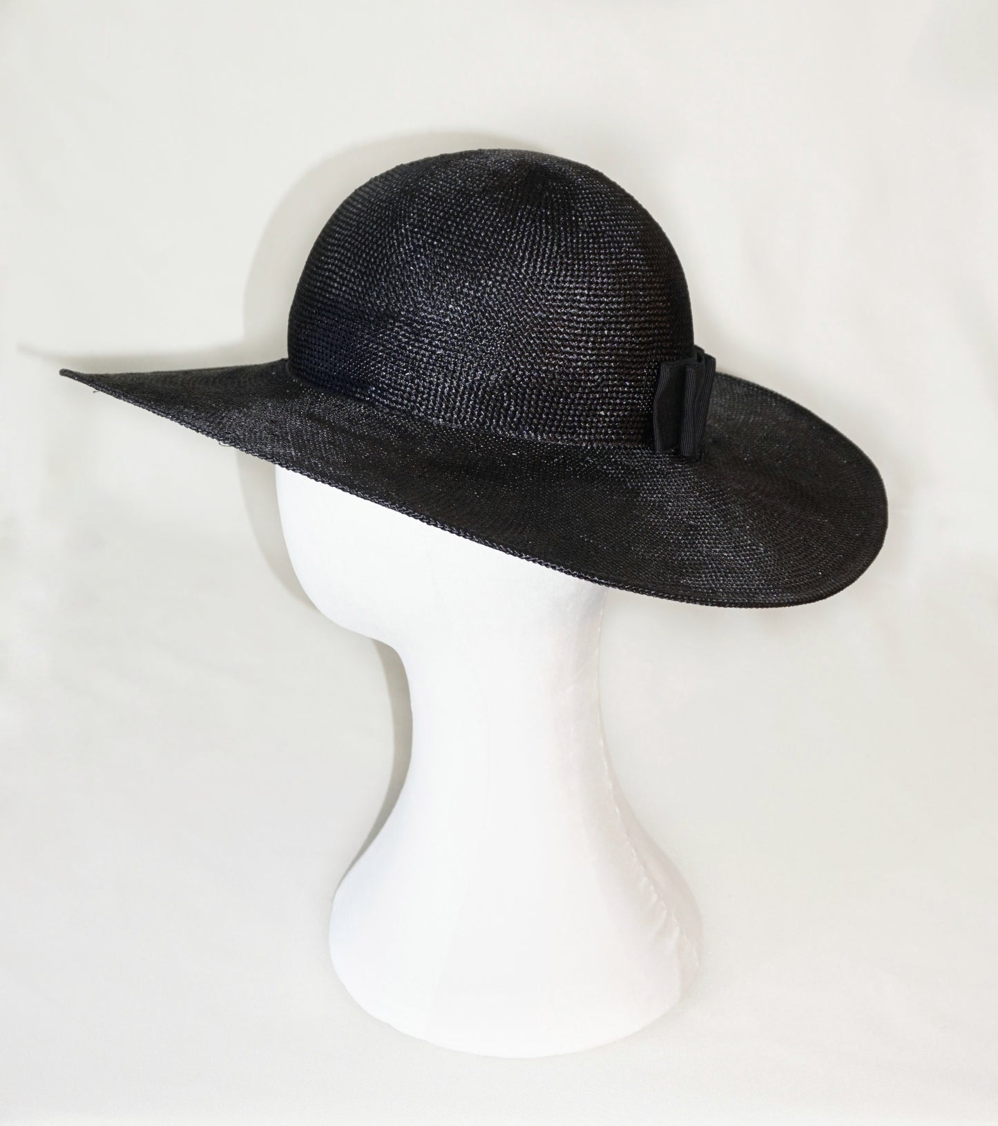 The handcrafted Auréole Élégante wide-brim visca straw hat in black, featuring a tailored black Italian cotton grosgrain bow and the signature 14ct gold-plated sterling silver lyre jewel hidden on the right back side, displayed on a white mannequin stand — left side view. Bespoke Couture Millinery by Anna Xaniotaki.