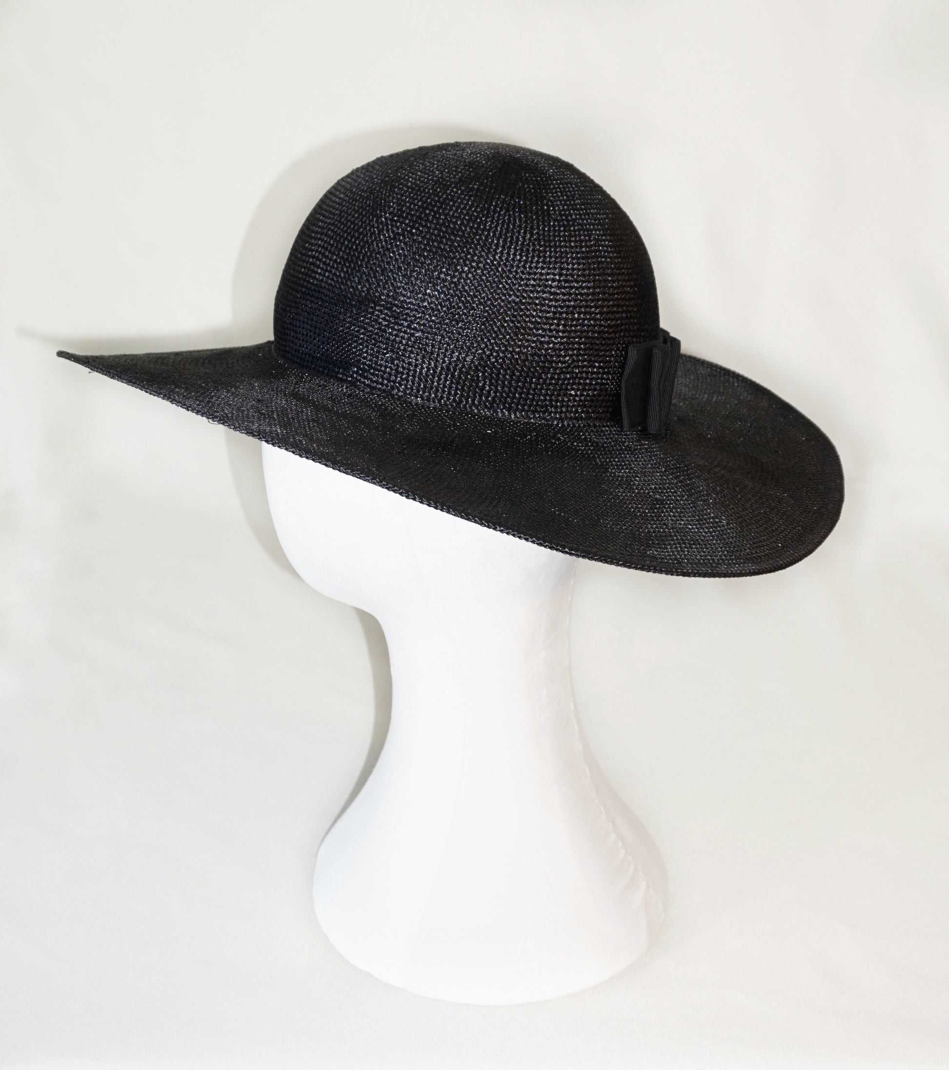 The handcrafted Auréole Élégante wide-brim visca straw hat in black, featuring a tailored black Italian cotton grosgrain bow and the signature 14ct gold-plated sterling silver lyre jewel hidden on the right back side, displayed on a white mannequin stand — left side view. Bespoke Couture Millinery by Anna Xaniotaki.