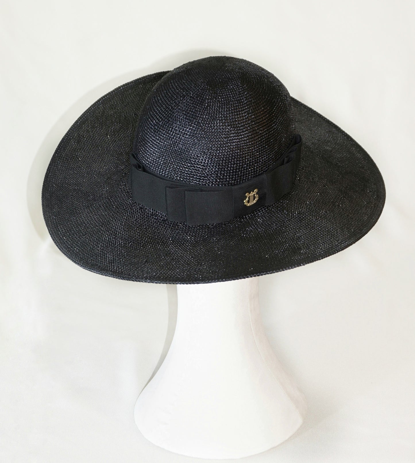 The handcrafted Auréole Élégante wide-brim visca straw hat in black, featuring a tailored black Italian cotton grosgrain bow and the signature 14ct gold-plated sterling silver lyre jewel, displayed on a white mannequin stand — high-angle three-quarter back left view. Bespoke Couture Millinery by Anna Xaniotaki.