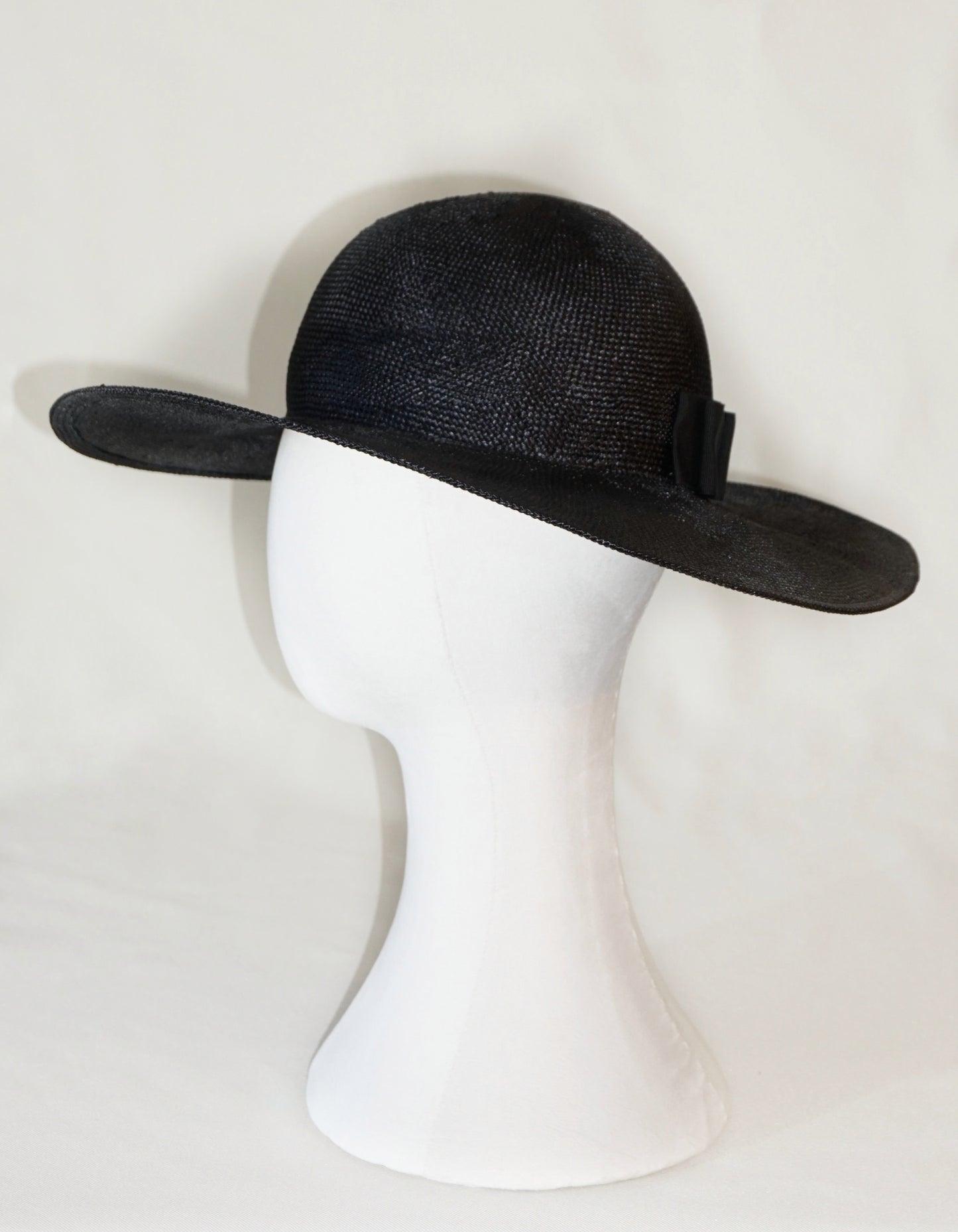 The handcrafted Auréole Élégante wide-brim visca straw hat in black, featuring a tailored black Italian cotton grosgrain bow and the signature 14ct gold-plated sterling silver lyre jewel hidden on the right back side, displayed on a white mannequin stand — left side view. Bespoke Couture Millinery by Anna Xaniotaki.