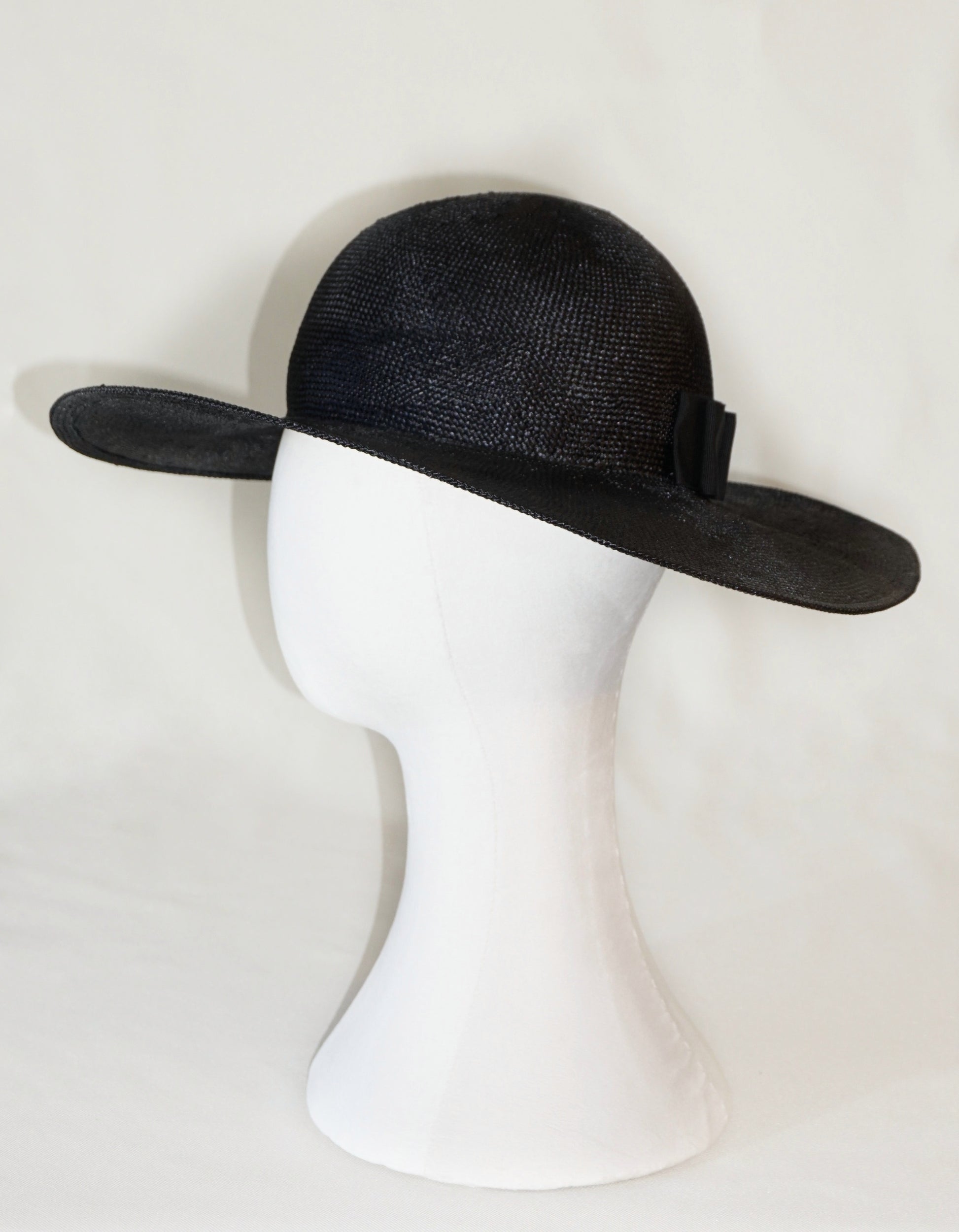 The handcrafted Auréole Élégante wide-brim visca straw hat in black, featuring a tailored black Italian cotton grosgrain bow and the signature 14ct gold-plated sterling silver lyre jewel hidden on the right back side, displayed on a white mannequin stand — left side view. Bespoke Couture Millinery by Anna Xaniotaki.
