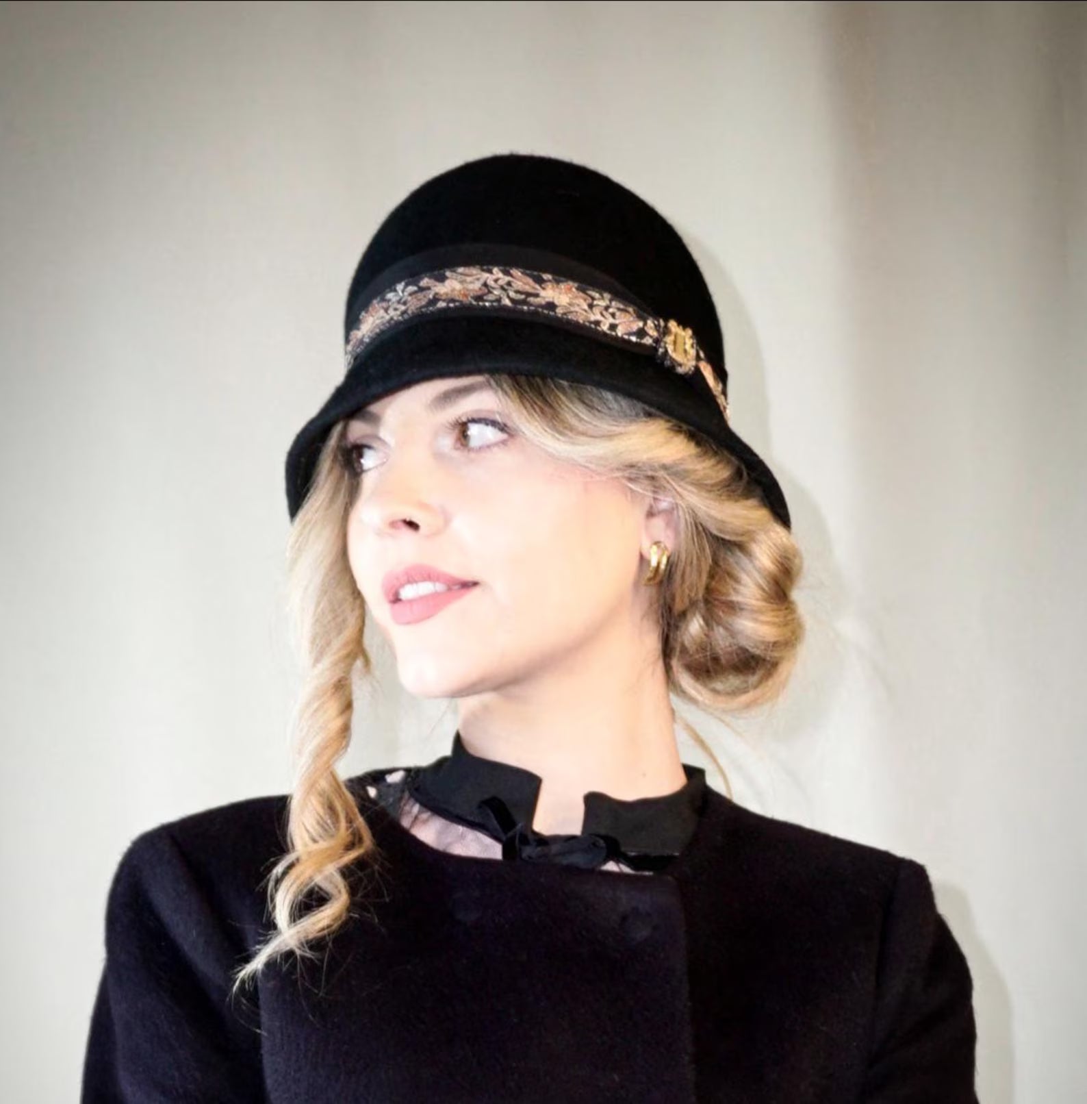 Woman wearing the Vintage Whisper black wool felt cloche hat with a gold lyre emblem, featuring an Italian black grosgrain ribbon and a floral woven ribbon. Three-quarter right side front view, handcrafted by Anna Xaniotaki.