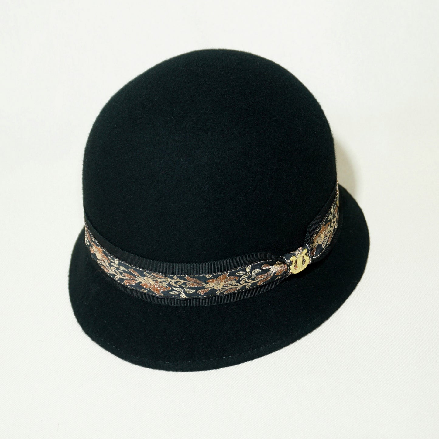 Vintage Whisper black wool felt cloche hat with a gold lyre emblem, featuring an Italian black grosgrain ribbon and a floral woven ribbon. Three-quarter top right side front view, handcrafted by Anna Xaniotaki