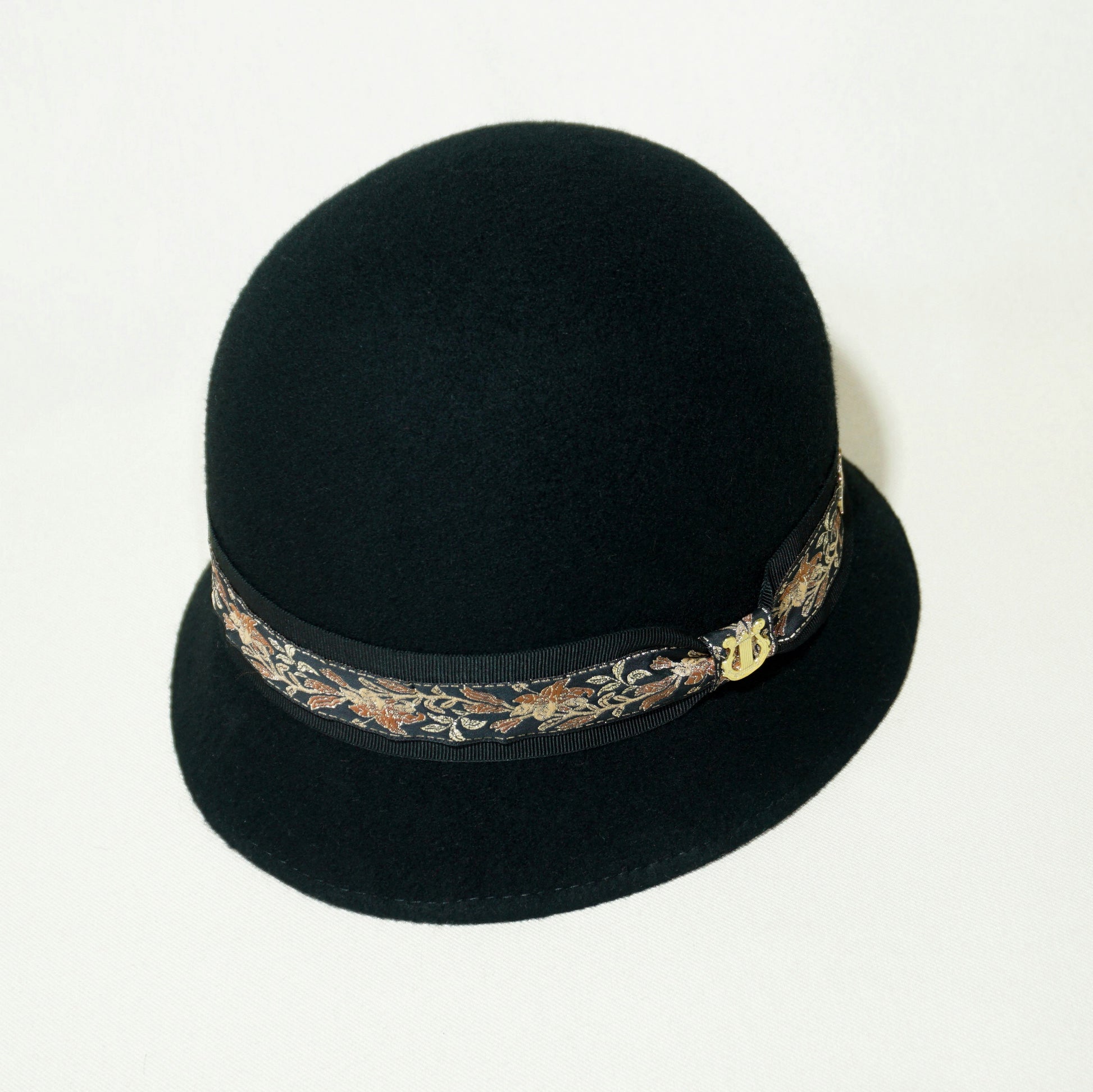 Vintage Whisper black wool felt cloche hat with a gold lyre emblem, featuring an Italian black grosgrain ribbon and a floral woven ribbon. Three-quarter top right side front view, handcrafted by Anna Xaniotaki