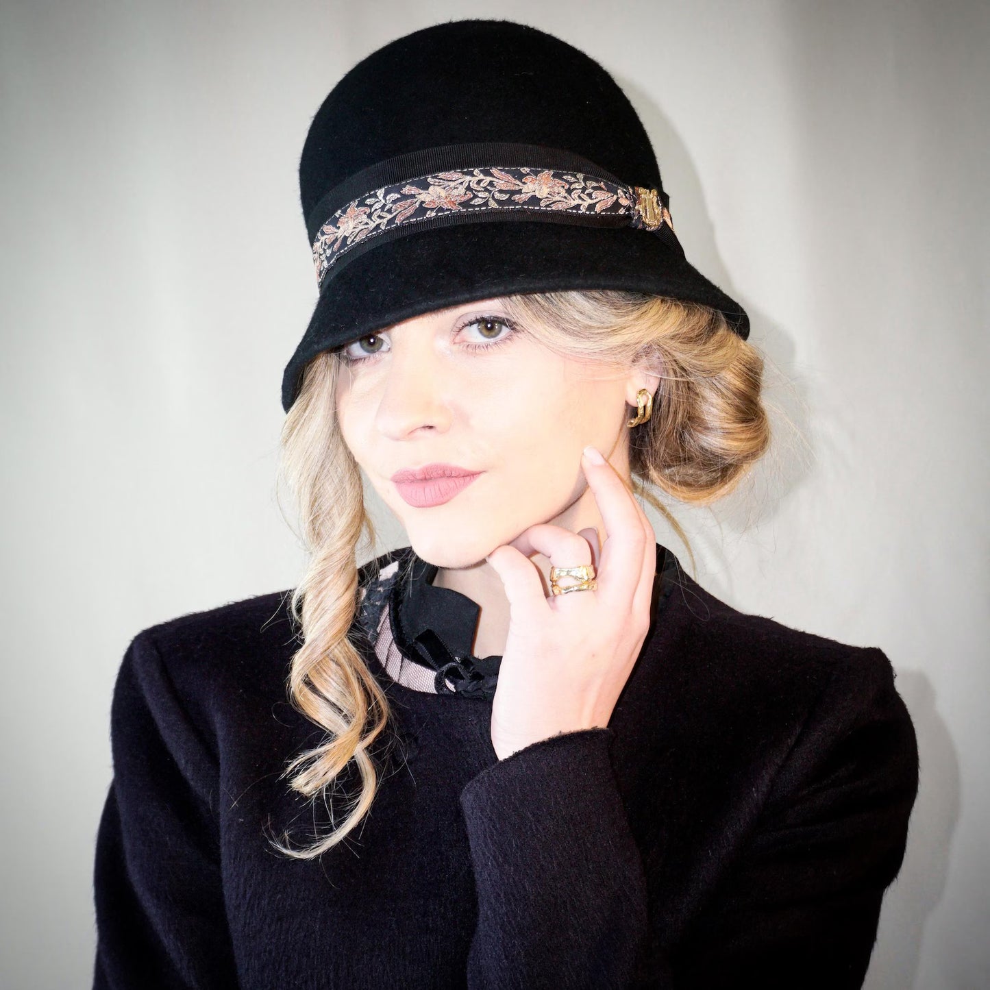 Woman wearing the Vintage Whisper black wool felt cloche hat with a gold lyre emblem, featuring an itallian black grosgrain ribbon and a floral woven ribbon. Three-quarter right side front view, handcrafted by Anna Xaniotaki.