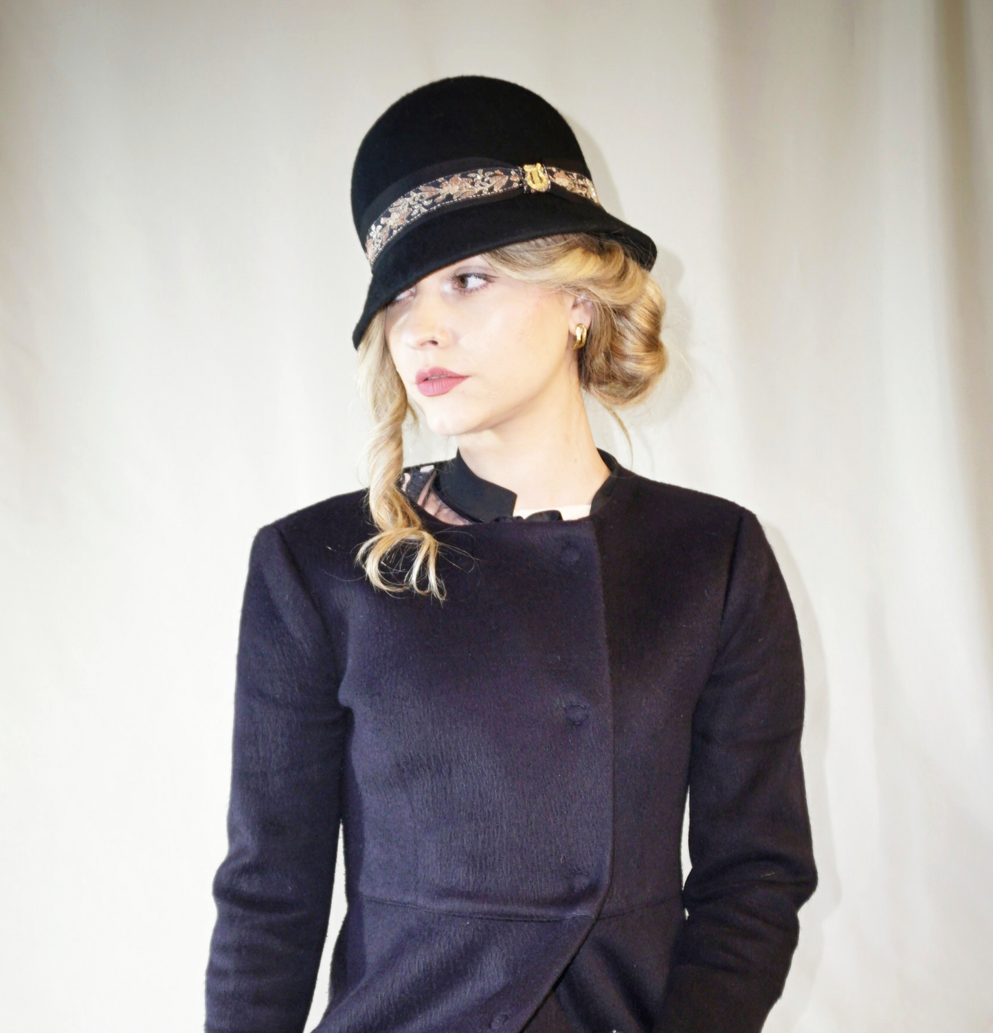 Woman wearing the Vintage Whisper black wool felt cloche hat with a gold lyre emblem, featuring an Italian black grosgrain ribbon and a floral woven ribbon. Three-quarter right side front view, handcrafted by Anna Xaniotaki.